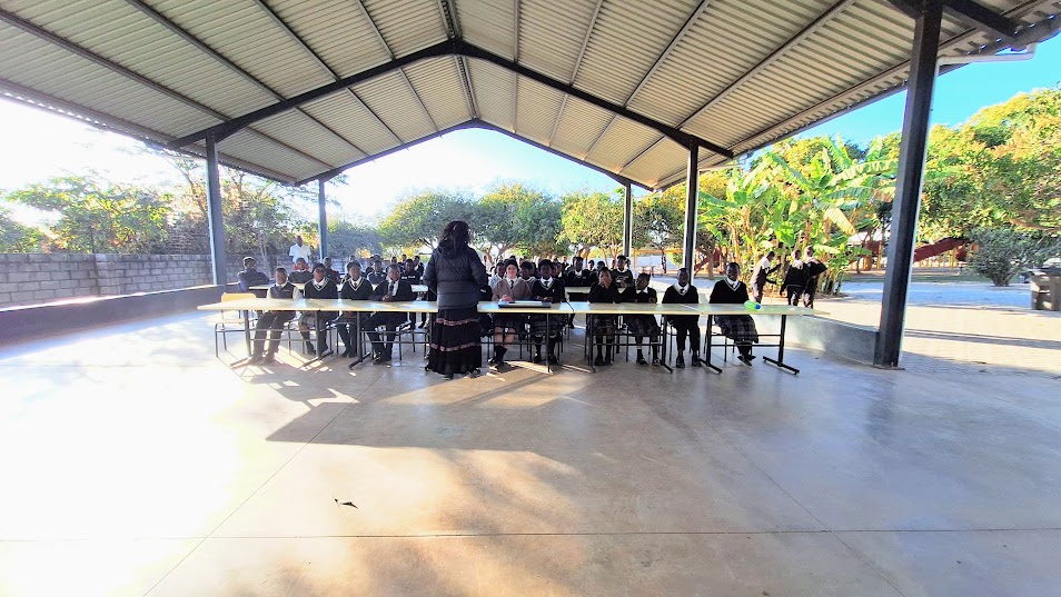 Secondary school students seated at long tables under an open-sided shelter, listening to a team member